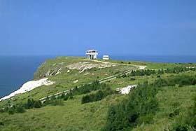 青空の下、奥に海が見える位置から筆ん崎の崖上にある緑広がるマハナ展望台を撮影した写真